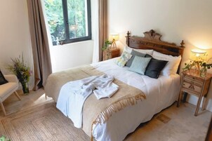 Una cama bien hecha con cabecera de madera, dos mesitas de noche con lámparas, una silla y una ventana con cortinas.