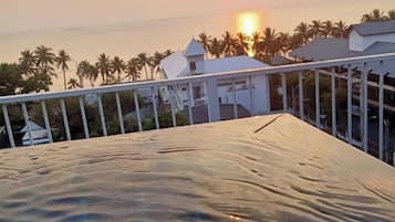 A balcony with a view of the sunset over the ocean, palm trees, and a white building with a sloped roof.