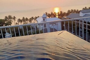 A balcony with a view of the sunset over the ocean, palm trees, and a white building with a sloped roof. - Baan Chid Haad (Chon Buri)