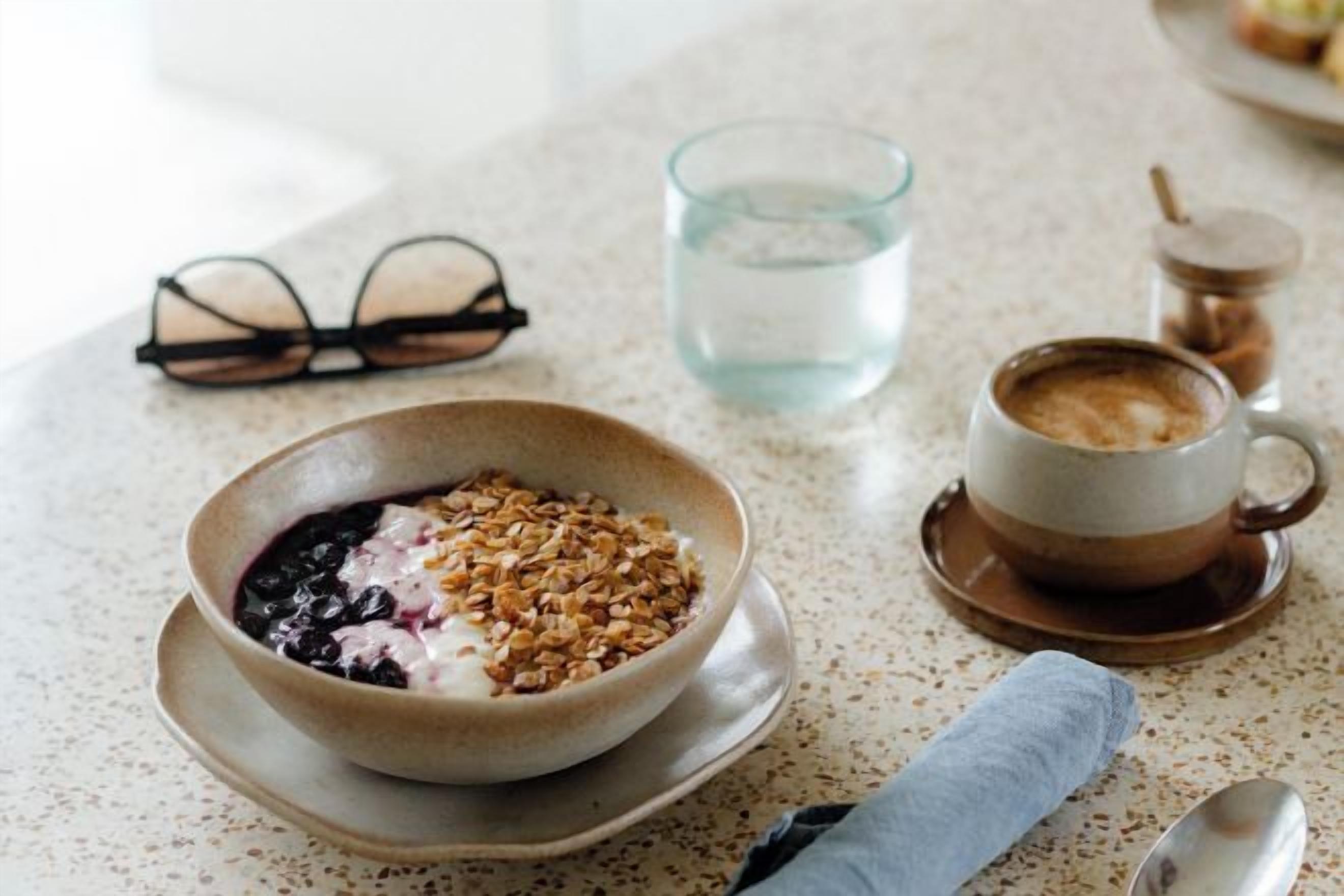Un desayuno con un tazón de cereal, una taza de café, un vaso de agua y un par de lentes sobre una encimera con motas.