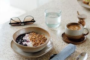 Une table mise pour le petit-déjeuner avec un bol de céréales, une tasse de café, un verre d’eau et une paire de lunettes posées sur un comptoir à motifs.