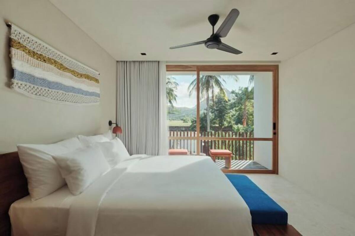 Ein Schlafzimmer mit Bett, Deckenventilator, Blick ins Freie und einem Balkon mit Holzgeländer.