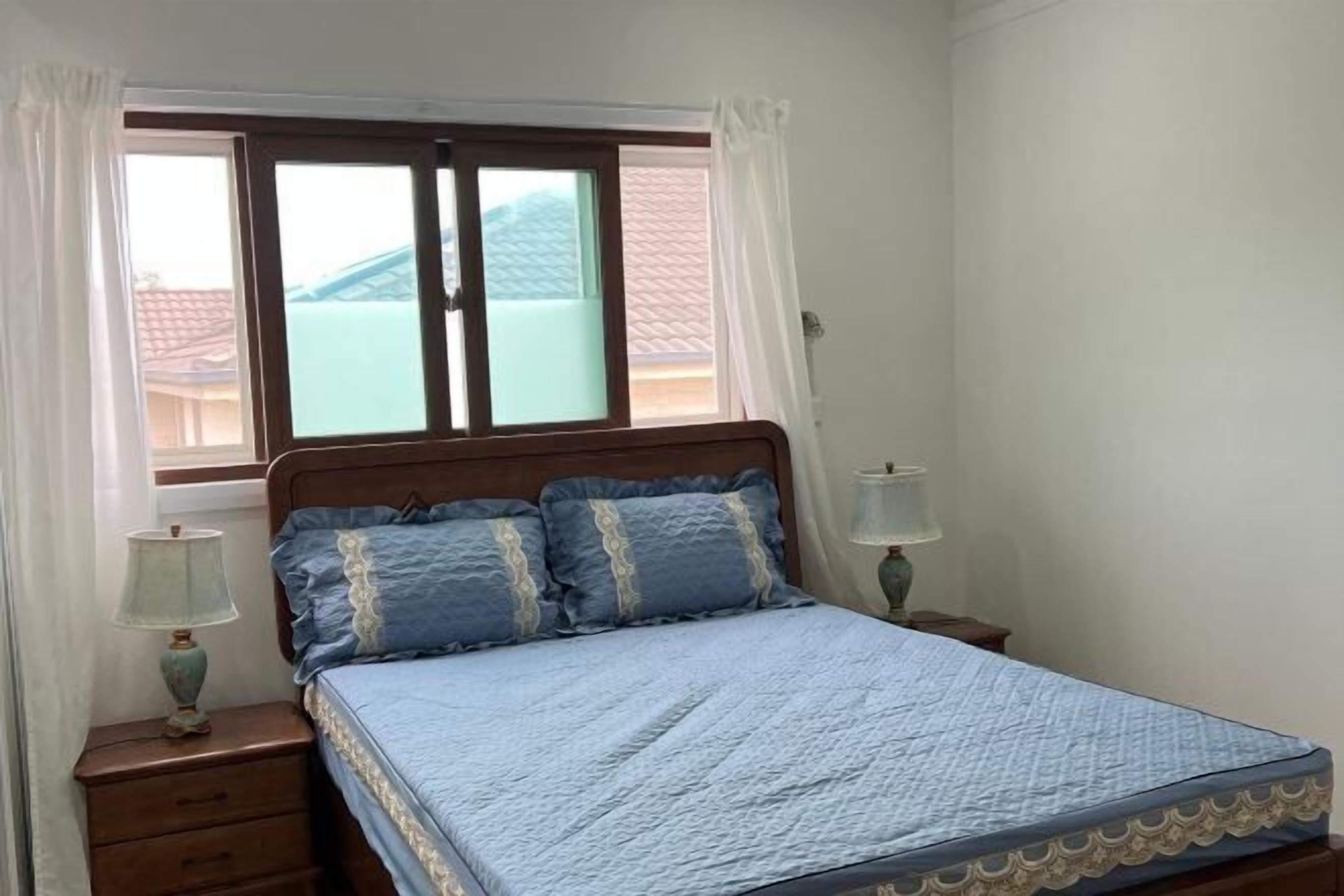Een slaapkamer met een bed, twee bedlampjes, een raam met gordijnen en uitzicht op een gebouw.