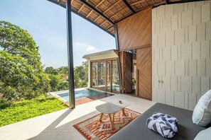 A modern room with a large glass door leading to a pool, a sofa with a patterned rug, and a view of lush greenery. - BAz Penthouse (Bali)