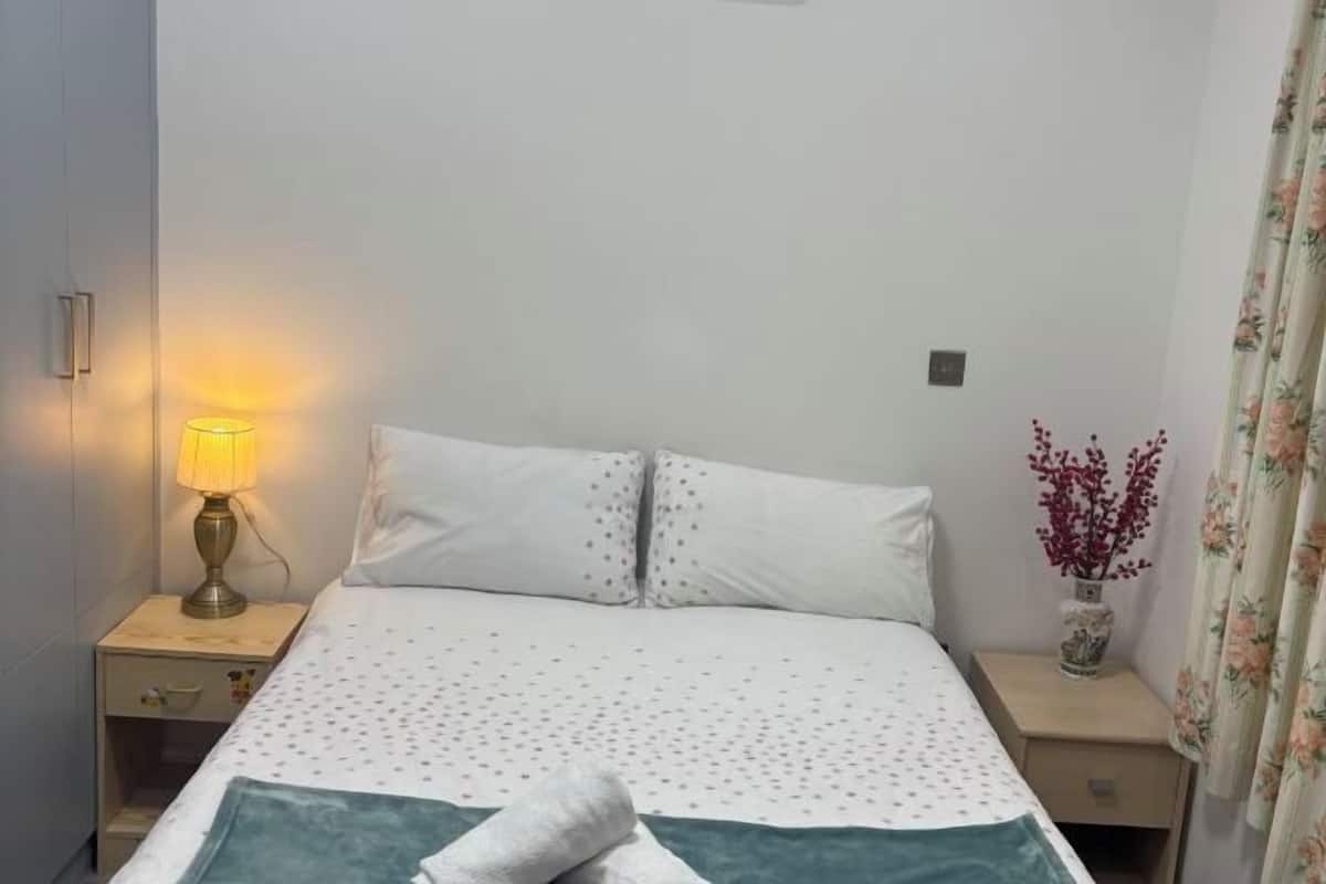 A neatly made bed with a white duvet and two pillows, a bedside lamp, a small vase with red flowers, and a floral curtain.