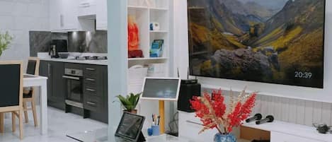 A modern kitchen with white cabinetry, a dining area with a glass table, and a large flat-screen TV mounted on the wall displaying a scenic landscape.