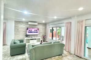 A modern living room with a flat-screen TV, green sofas, a white entertainment unit, and a large sliding glass door leading to an outdoor area.