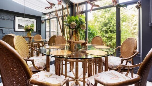 Una mesa de vidrio redonda rodeada de sillas de mimbre en una habitación con ventanas amplias y figuras decorativas de pájaros.