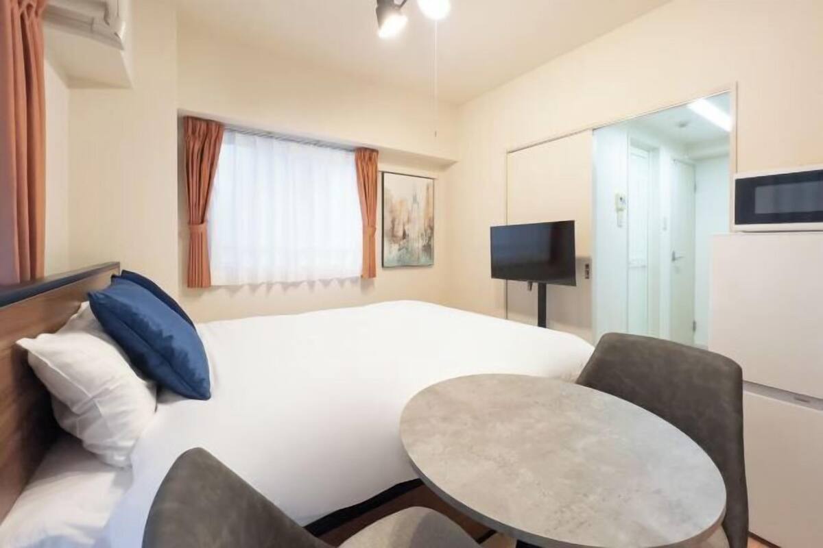 Une chambre d’hôtel avec un lit, une petite table ronde, une chaise, un téléviseur et une fenêtre avec des rideaux.