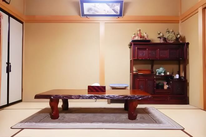 Une pièce de style japonais traditionnel, avec un sol recouvert de tatamis, une basse table en bois, un téléviseur fixé au mur et un meuble en bois contenant des objets décoratifs.