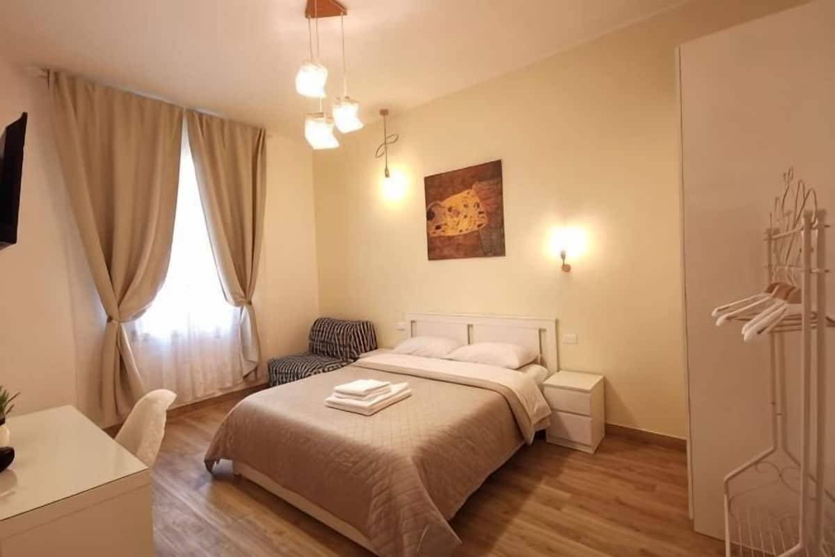 A bedroom with a bed, bedside tables, a chair, a desk, a wardrobe, and a hanging rack.