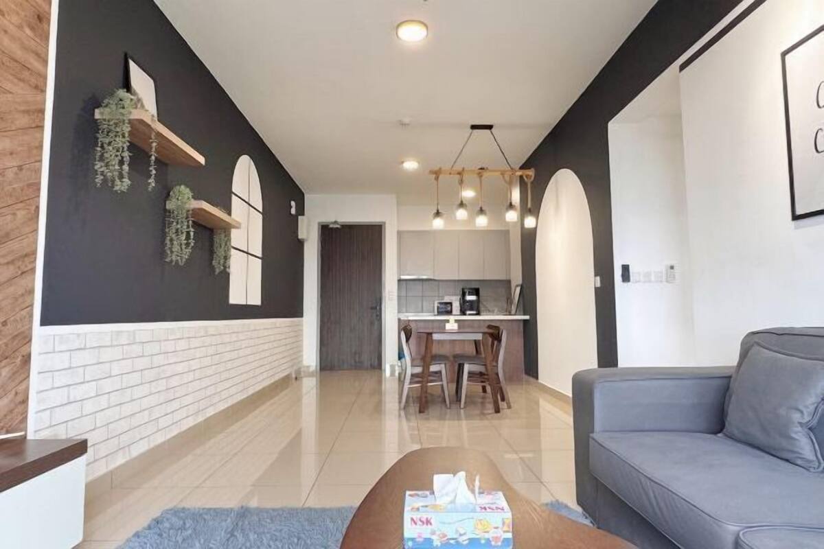 A modern living room with a sofa, a coffee table with a box, a kitchen area with a dining table and chairs, and a hallway with a door.