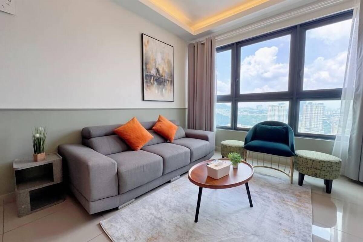 A modern living room with a grey sofa, orange cushions, a round coffee table, and a view of a cityscape through large windows.