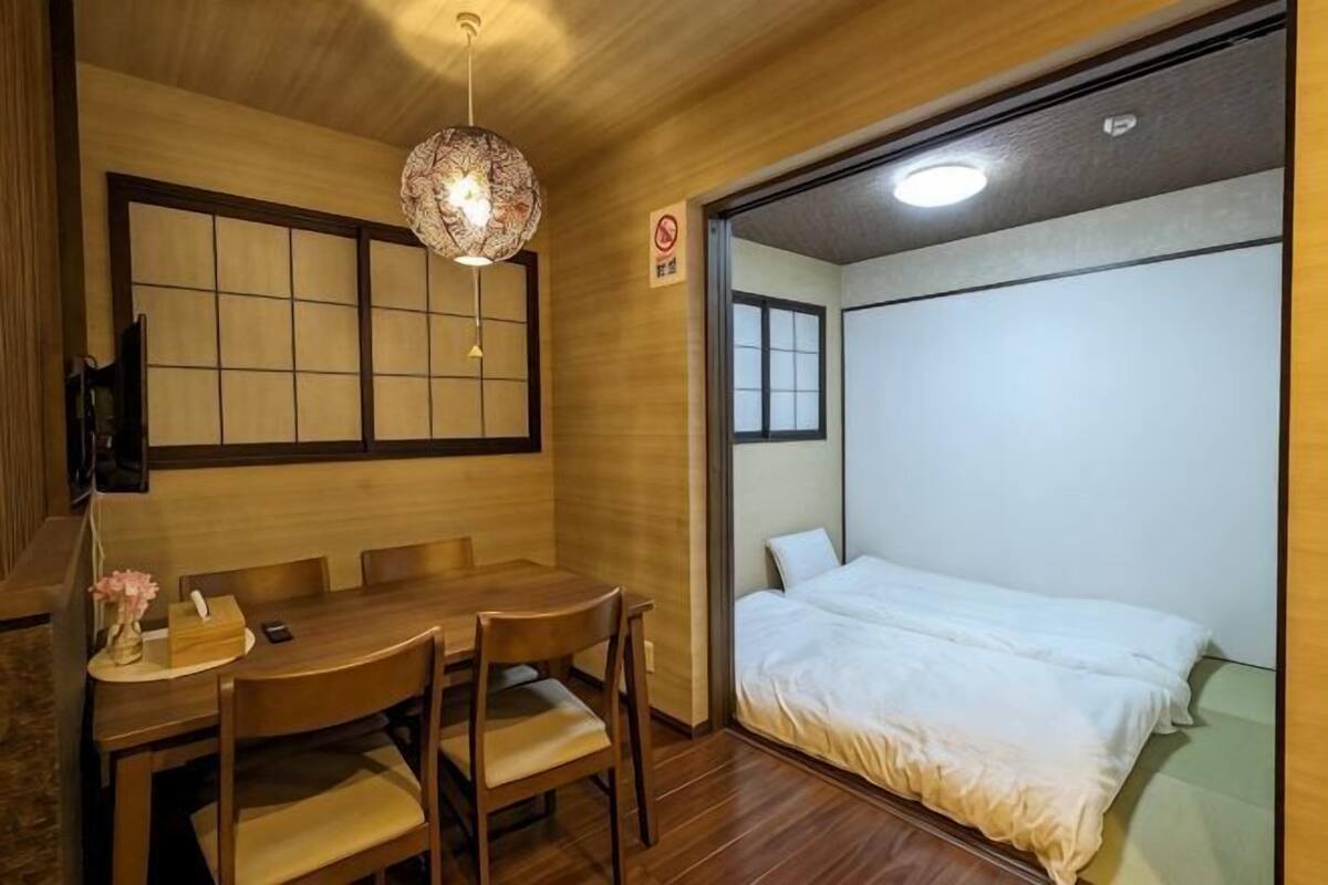 A room with a wooden dining table, chairs, a hanging light fixture, a bed with white bedding, and a sliding door.