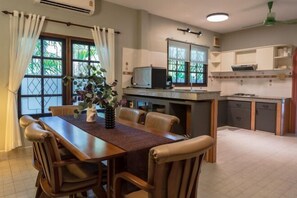 Een eetgedeelte met een houten tafel en stoelen, een kleine keuken met een magnetron, en een raam met gordijnen.