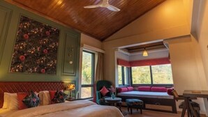 A bedroom with a bed, a seating area with a bench, a chair, and a table. There are windows providing natural light and a view of greenery outside. - Chakk 109 Mussoorie Foothills, Dehradun (Dehradun)