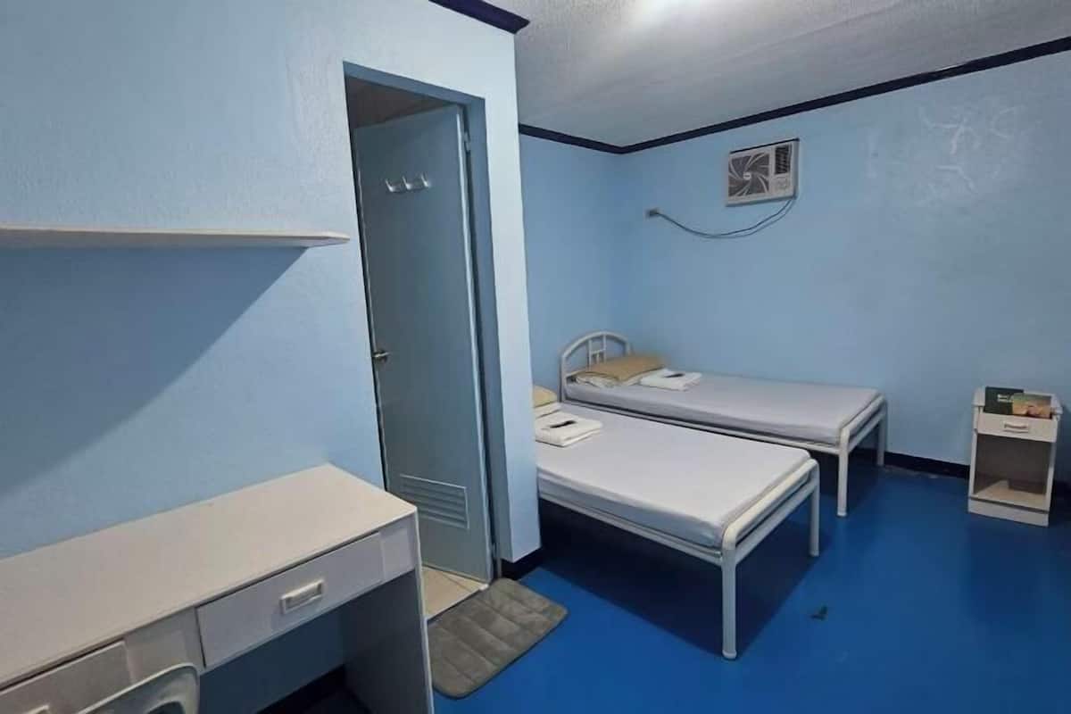 A small, single-room accommodation with a desk, two single beds, a door, and a small shelf.