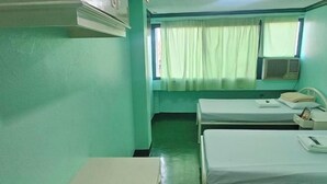 A room with two beds, a table, a chair, a window with curtains, and an air conditioning unit. - Green Mellow Court Capitol Avenue (Dipolog)