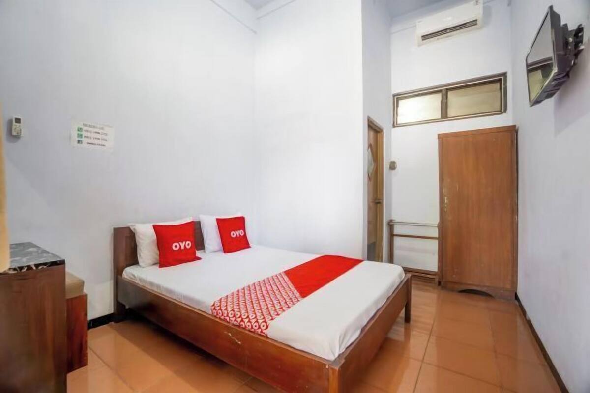 A hotel room with a bed, a wooden wardrobe, a small table, and a wall-mounted air conditioner.