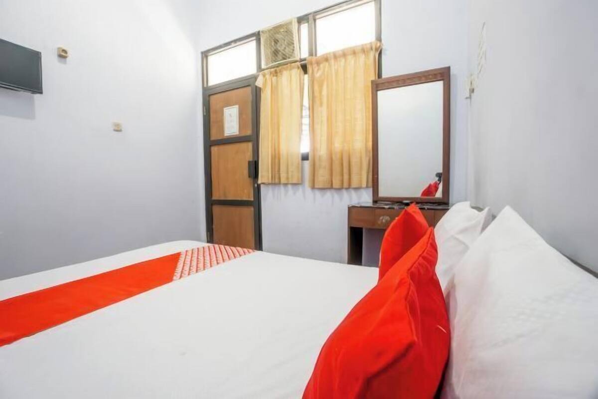 A hotel room with a bed, a wooden desk, a mirror, and a window with curtains.