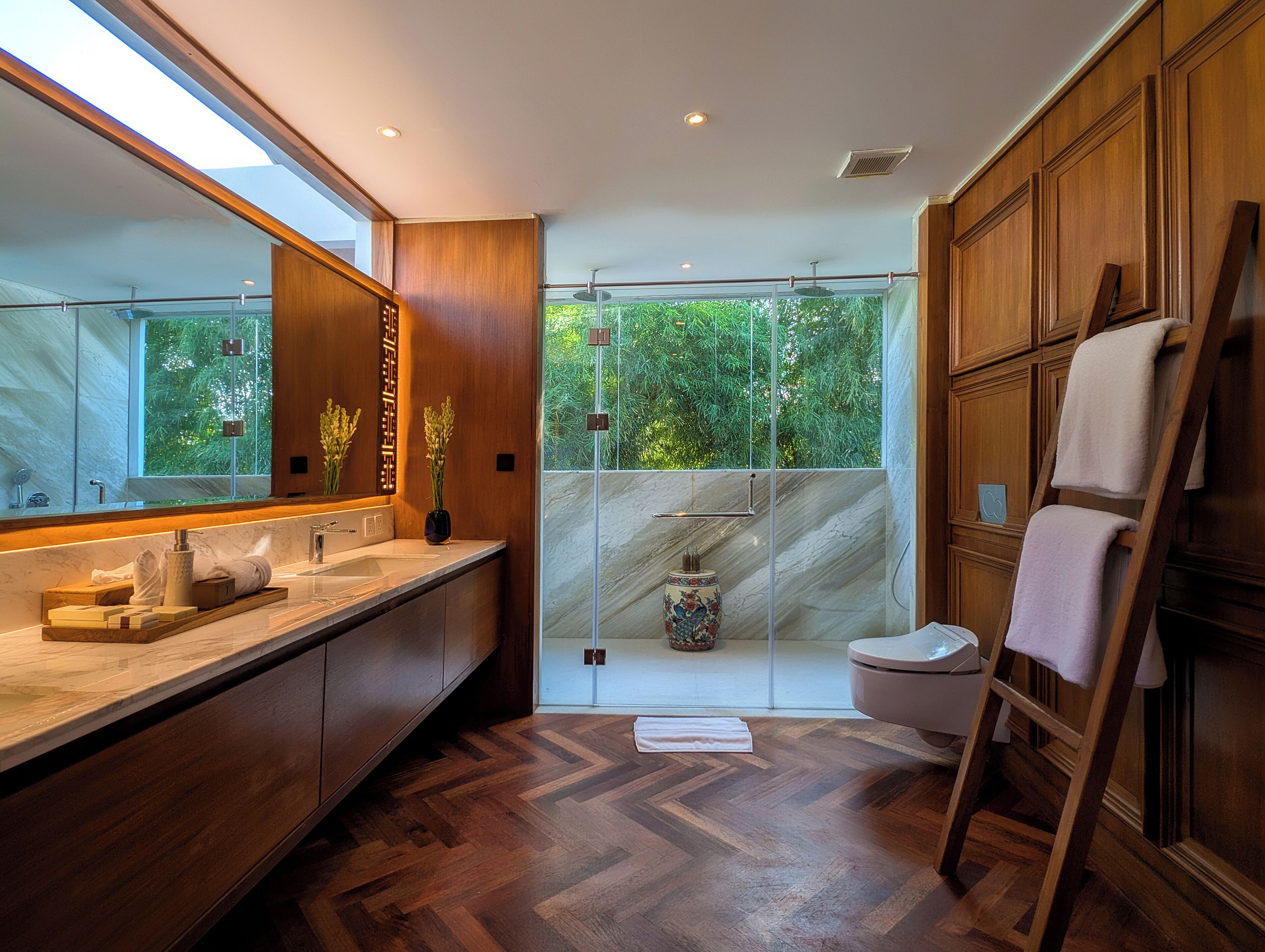 A modern bathroom with a large mirror, wooden cabinets, a marble countertop, and a view of greenery through a glass door.
