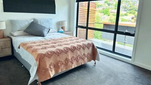 Une chambre à coucher avec un lit, des tables de chevet, une fenêtre donnant sur un paysage verdoyant et des bâtiments, et une tête de lit noire.