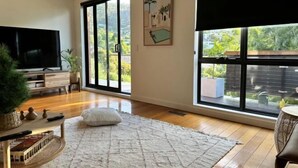 Un salon doté d’un grand téléviseur, d’un parquet, d’un tapis blanc et de grandes fenêtres offrant une vue sur la verdure.