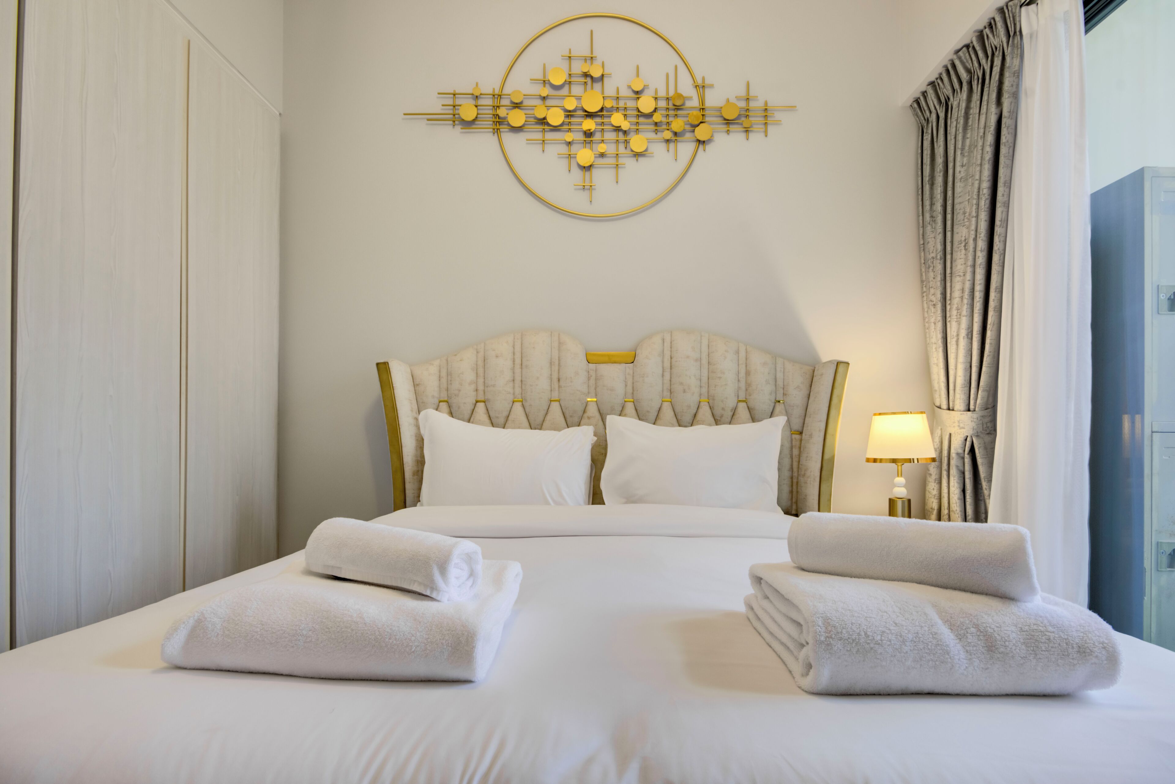 Ein Hotelzimmer mit einem großen Bett, zwei Kissen, einem Kopfteil, einem Wandbild, einer Lampe und einem Fenster mit Vorhängen.