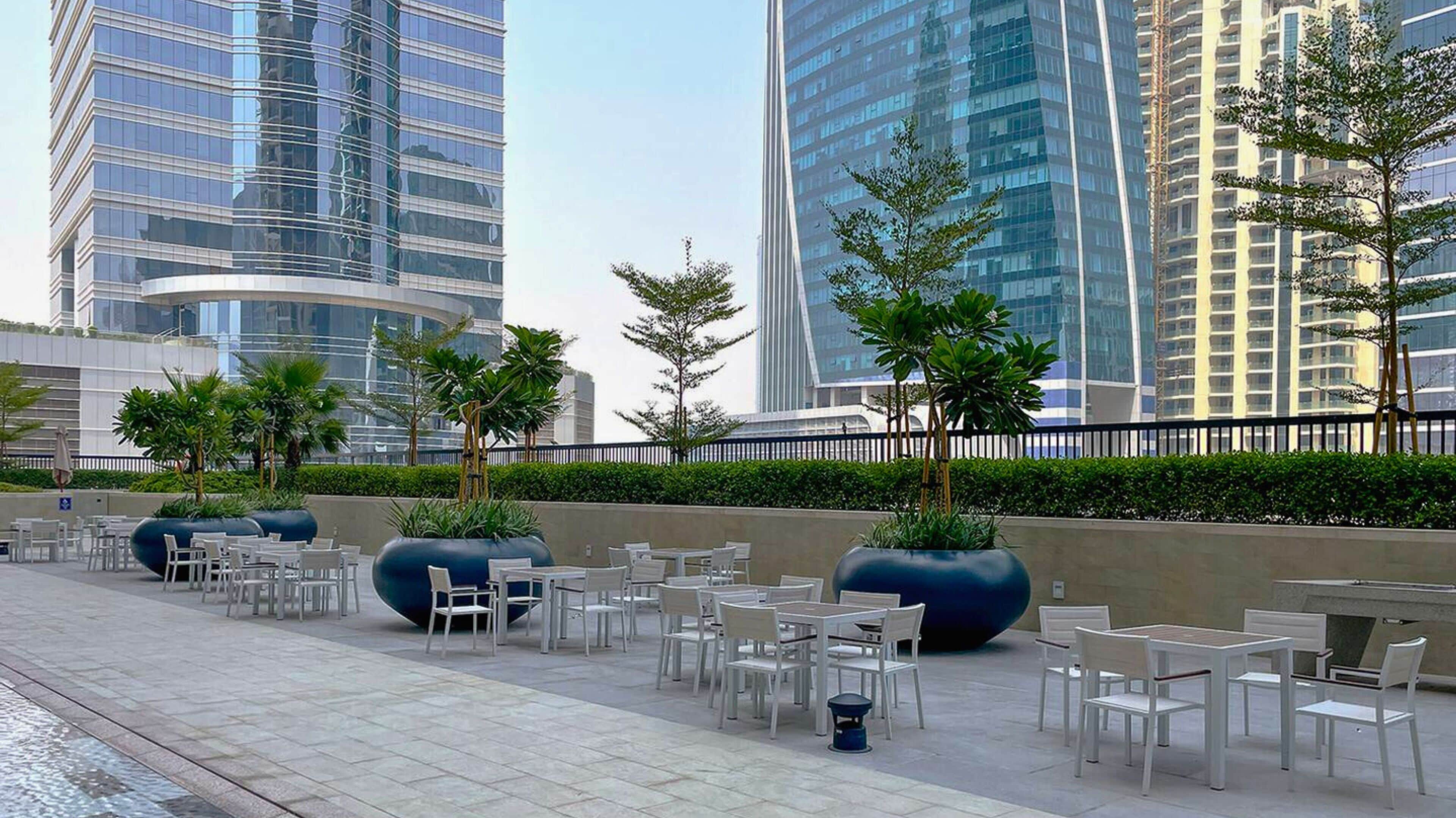 An outdoor seating area with tables and chairs, surrounded by tall buildings and greenery.
