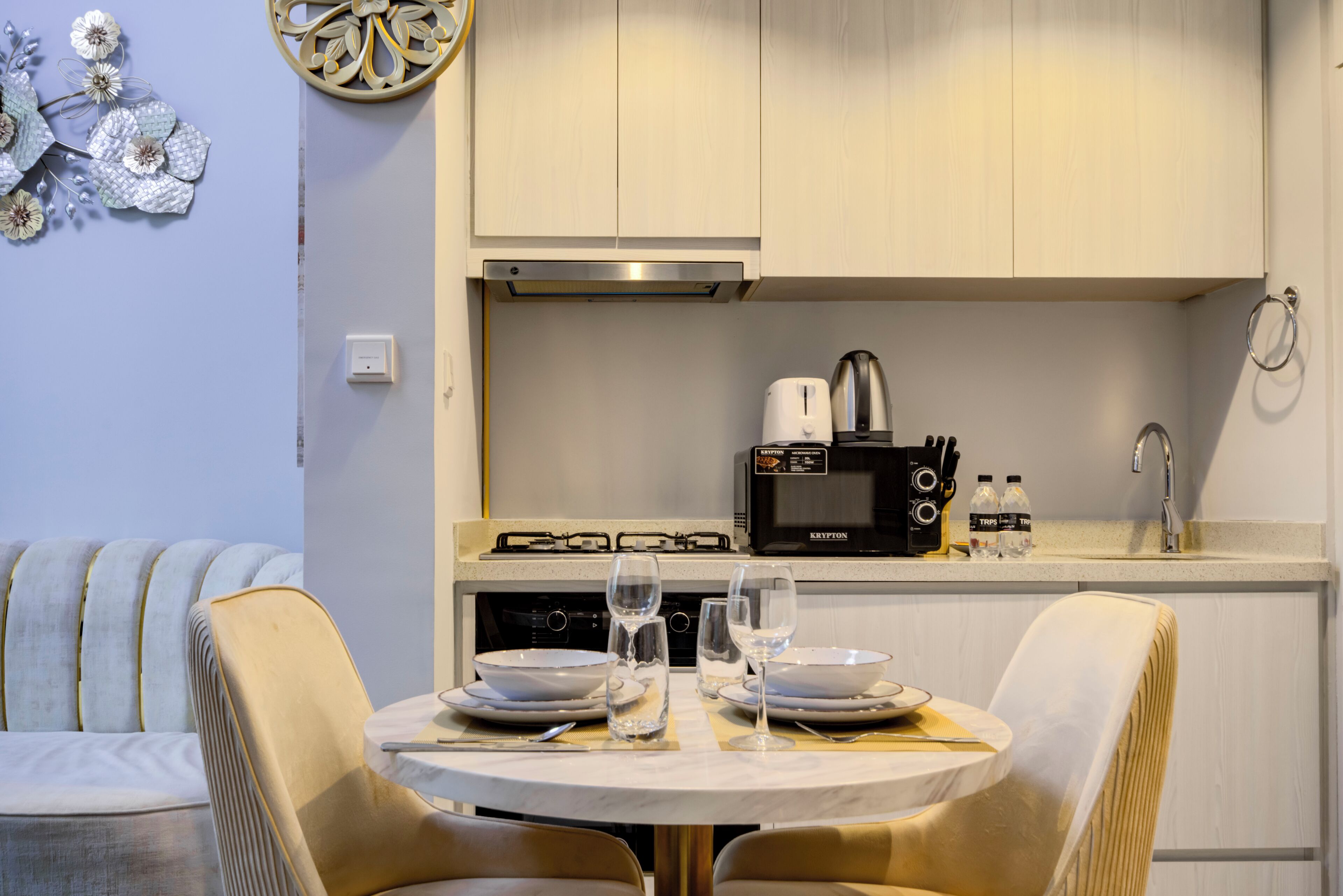 Uma cozinha compacta com micro-ondas, chaleira e uma pia pequena. Há uma mesa de jantar com cadeiras e um objeto decorativo na parede.