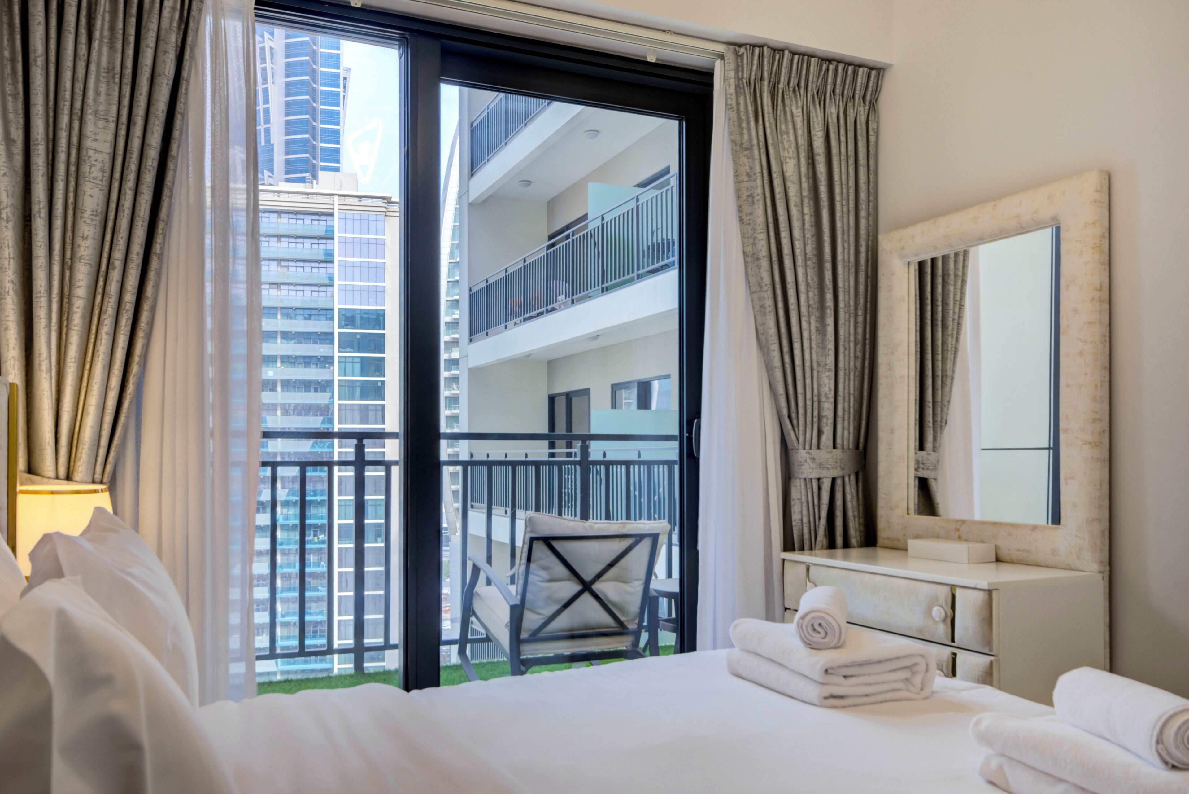 Un cuarto de hotel con cama, tocador, espejo y vistas a un balcón con una silla.