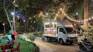 A food truck parked in a garden area with string lights, surrounded by greenery and a few people. - Keith Ratchaburi (Ban Pong)