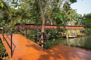 A wooden bridge over a pond with a railing, surrounded by greenery. - Keith Ratchaburi (Ban Pong)