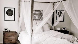 A four-poster canopy bed with a white bedspread, a wooden nightstand with a drawer, and two framed pictures on the wall. - Erykah Hotel Mityana Road (Mawokota)