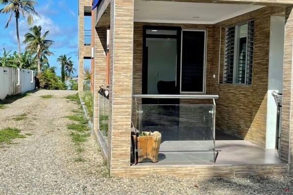 Um edifício moderno com entrada por porta de vidro, uma pequena área de estar externa e vista para palmeiras.