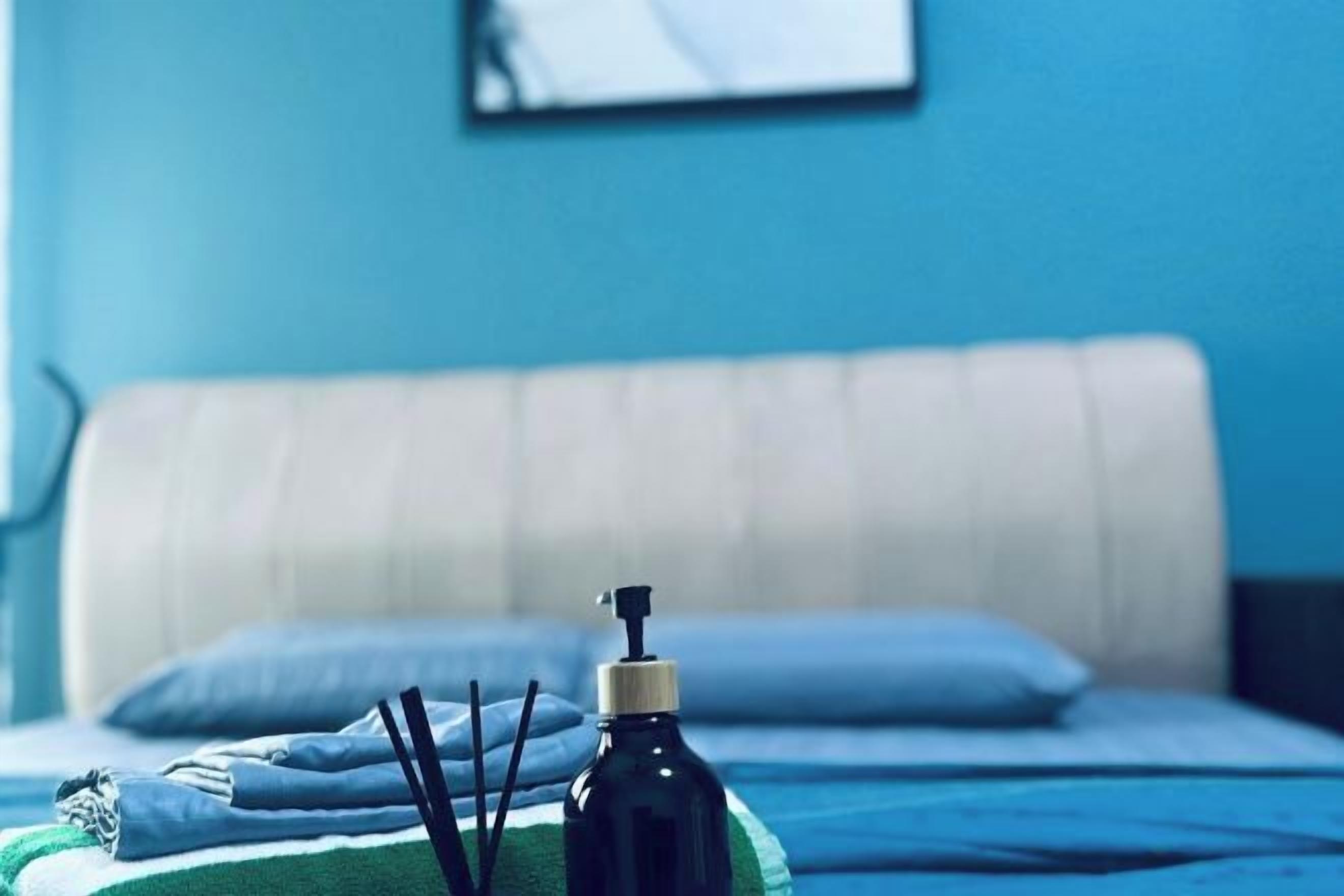 Una cama con una manta azul, un almohadón blanco y un frasco negro con dispensador.