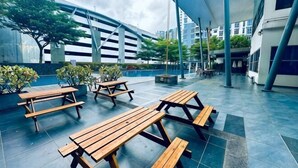 An outdoor patio with wooden picnic tables, a covered seating area, and a modern building in the background. - Johor Cozy Home 2 Rooms & Easy Parking Fast WiFi ! (Plentong)
