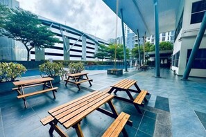 An outdoor patio with wooden picnic tables, a covered seating area, and a modern building in the background. - Johor Cozy Home 2 Rooms & Easy Parking Fast WiFi ! (Plentong)