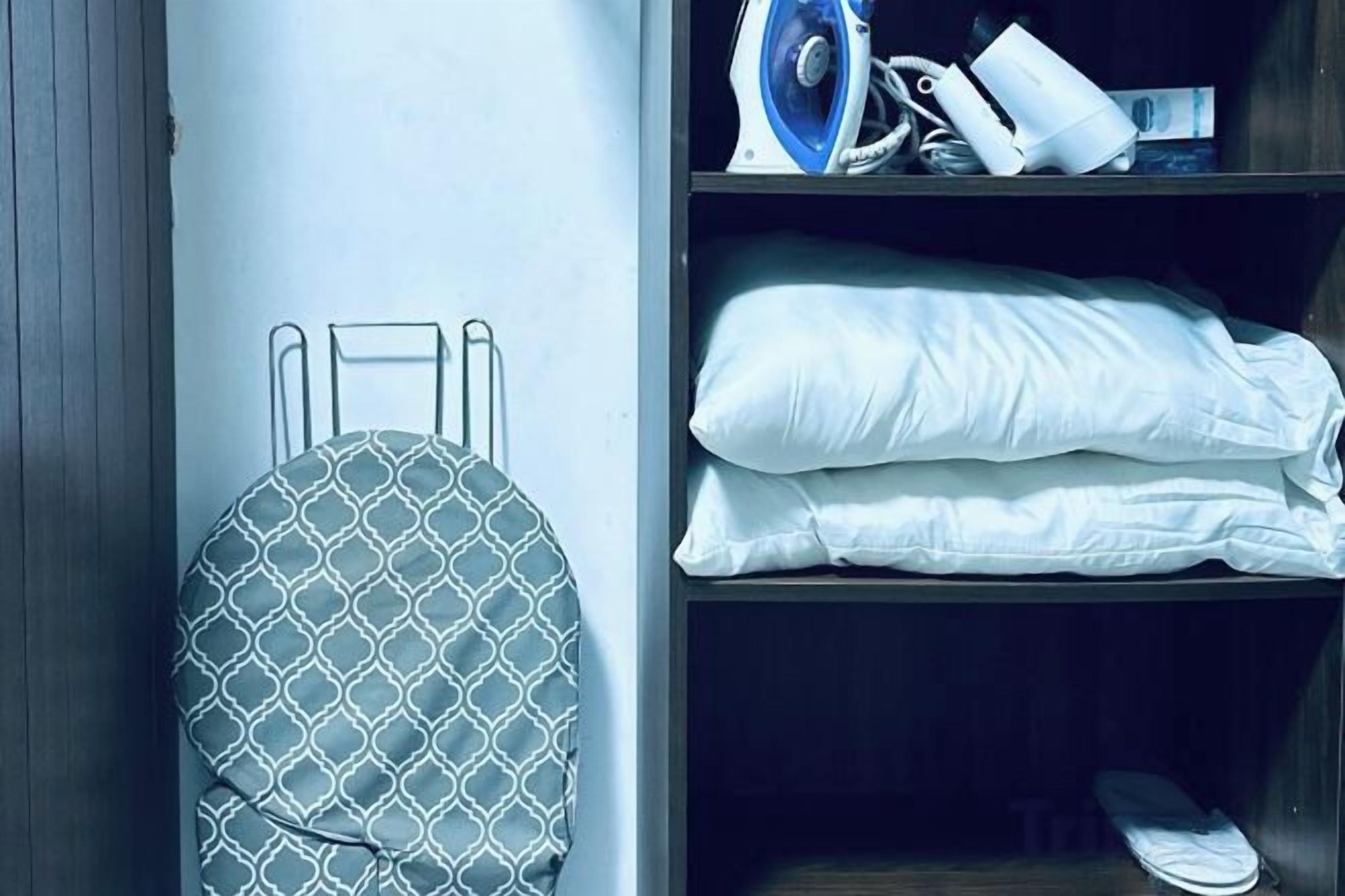 Une pièce bien rangée, avec un lit, un oreiller, une couverture pliée, une paire de chaussons et une planche à repasser fixée au mur.