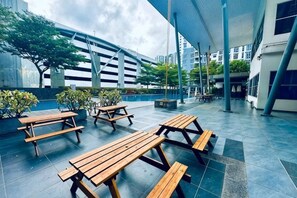 Un espacio al aire libre con mesas de picnic y bancos de madera, rodeado de edificios y follaje.