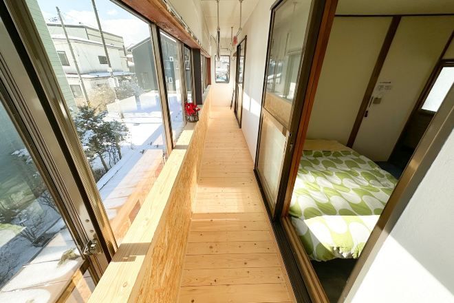 A narrow corridor with a wooden floor, large windows, and a bed with a green and white patterned bedspread.