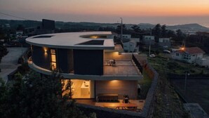 Une maison moderne à plusieurs étages, dotée d’un toit incurvé, d’un balcon et d’un petit espace extérieur aménagé pour s’asseoir.