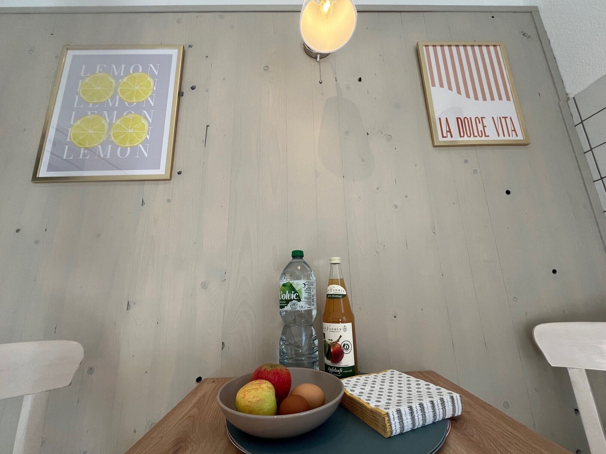 A room with a wooden table, a bowl of fruit, a bottle of water, a bottle of wine, and two framed pictures on the wall.