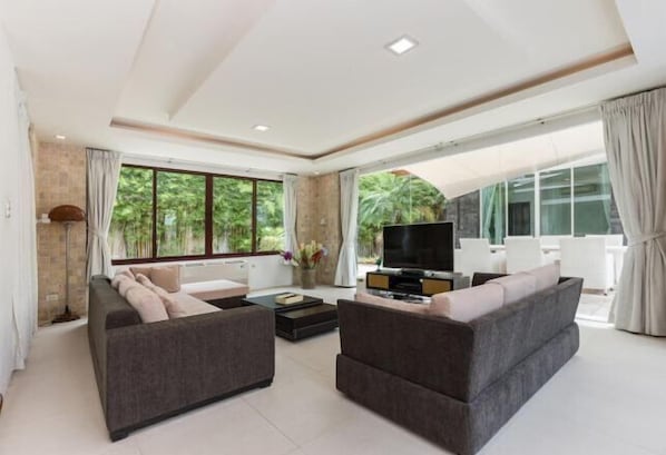 A modern living room with a sofa set, a flat-screen TV, and large windows offering a view of greenery.