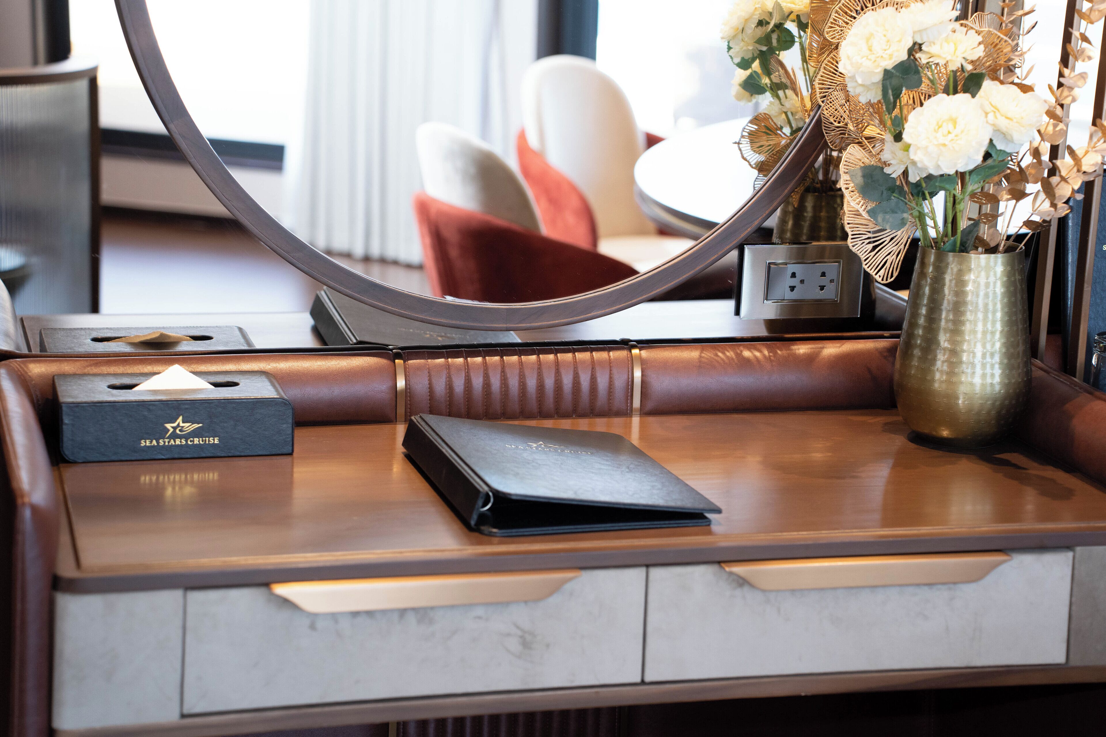A hotel desk with a laptop, a black box labeled "SEA STARS CRUISE," a vase with white flowers, and a mirror reflecting the room.
