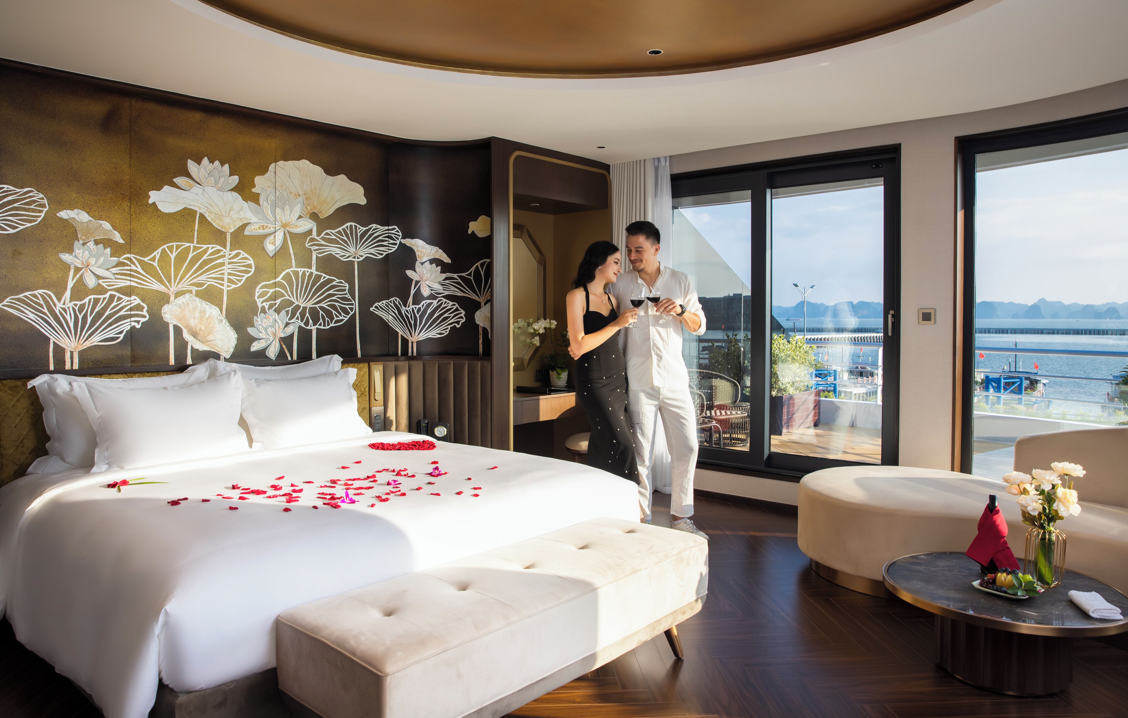 A modern hotel room with a large bed, a balcony with a view of the water, and a dining area with a table and chairs.