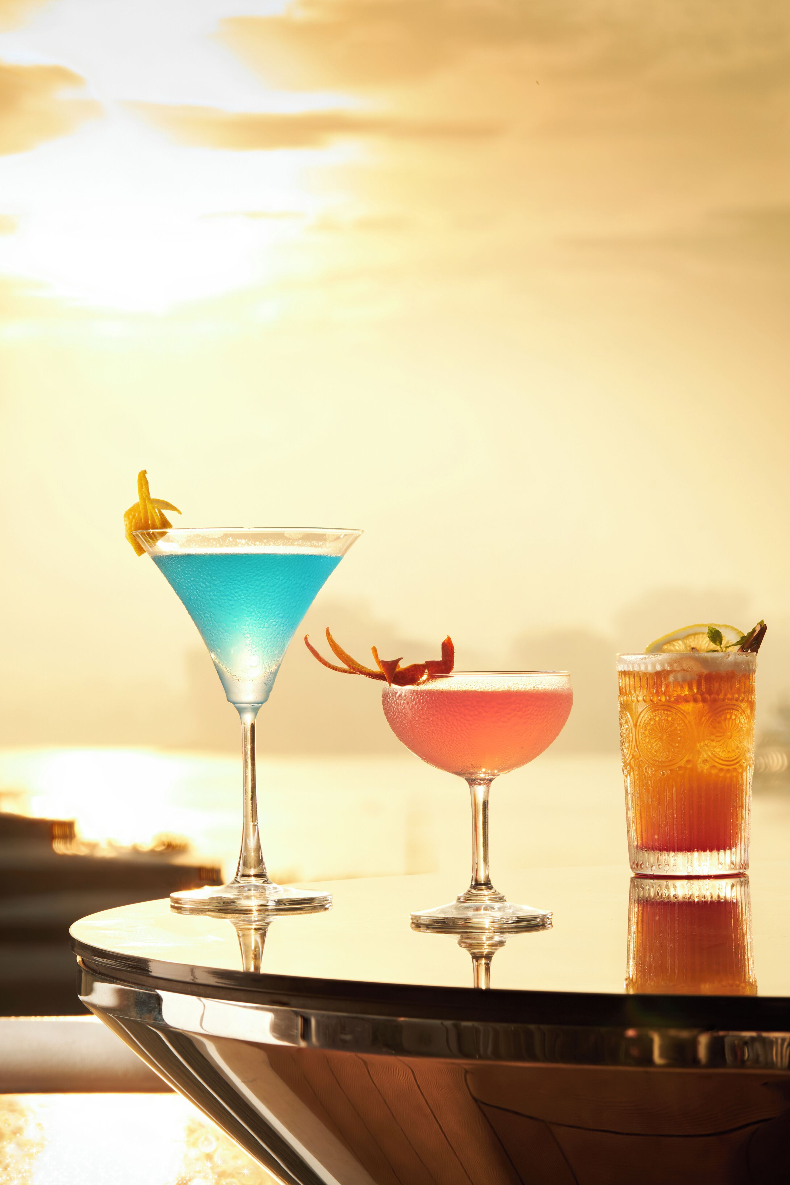 Three cocktails on a reflective surface with a sunset in the background.