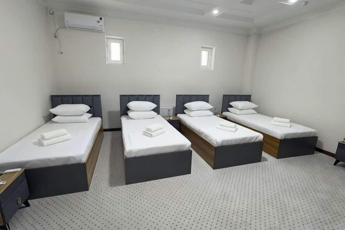Habitación de hotel con cuatro camas individuales, cada una con una almohada y una toalla. Se aprecia una ventana pequeña en la pared, y el suelo está alfombrado.