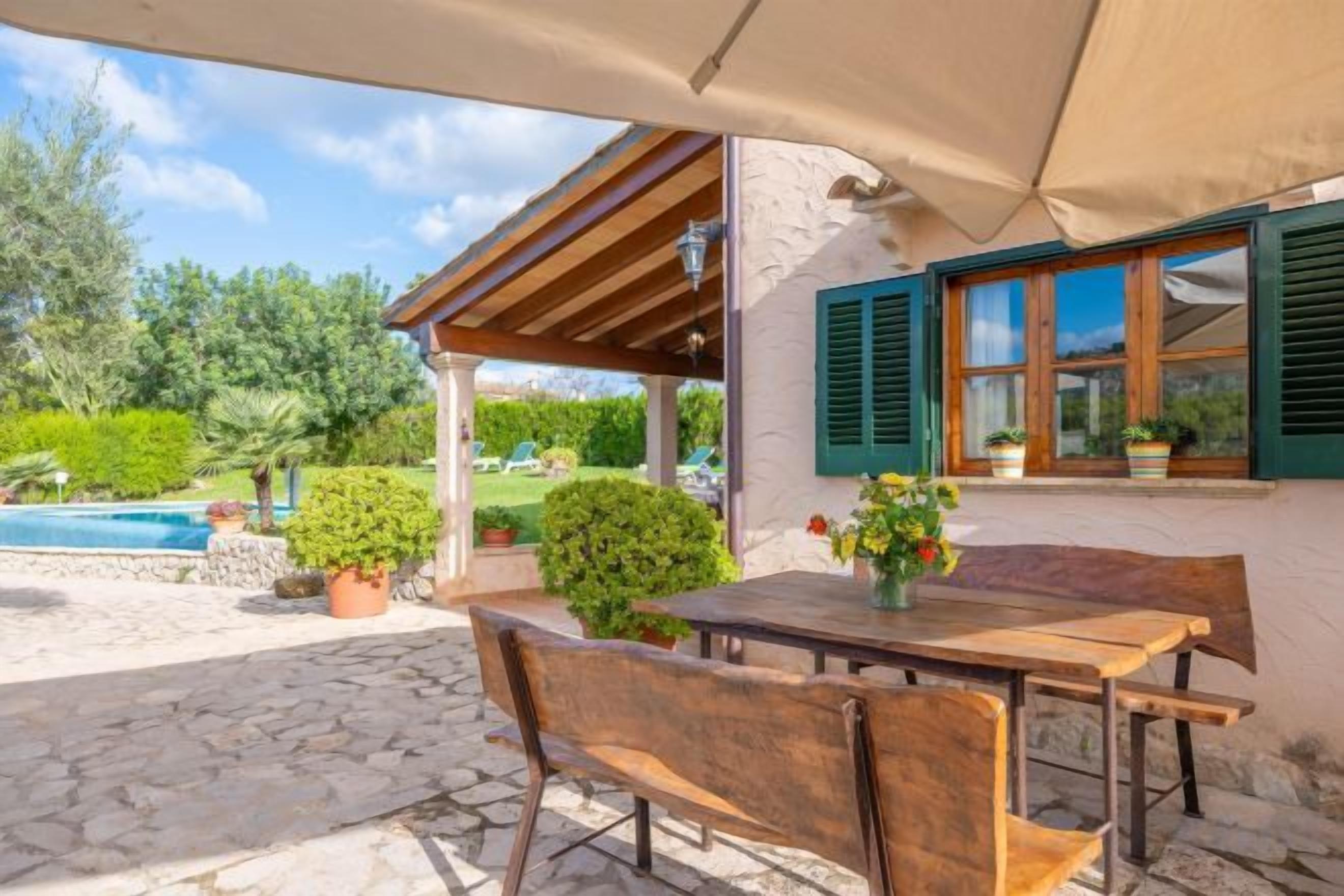 An outdoor patio with a wooden table and chairs, a covered area, and a view of a swimming pool and greenery.