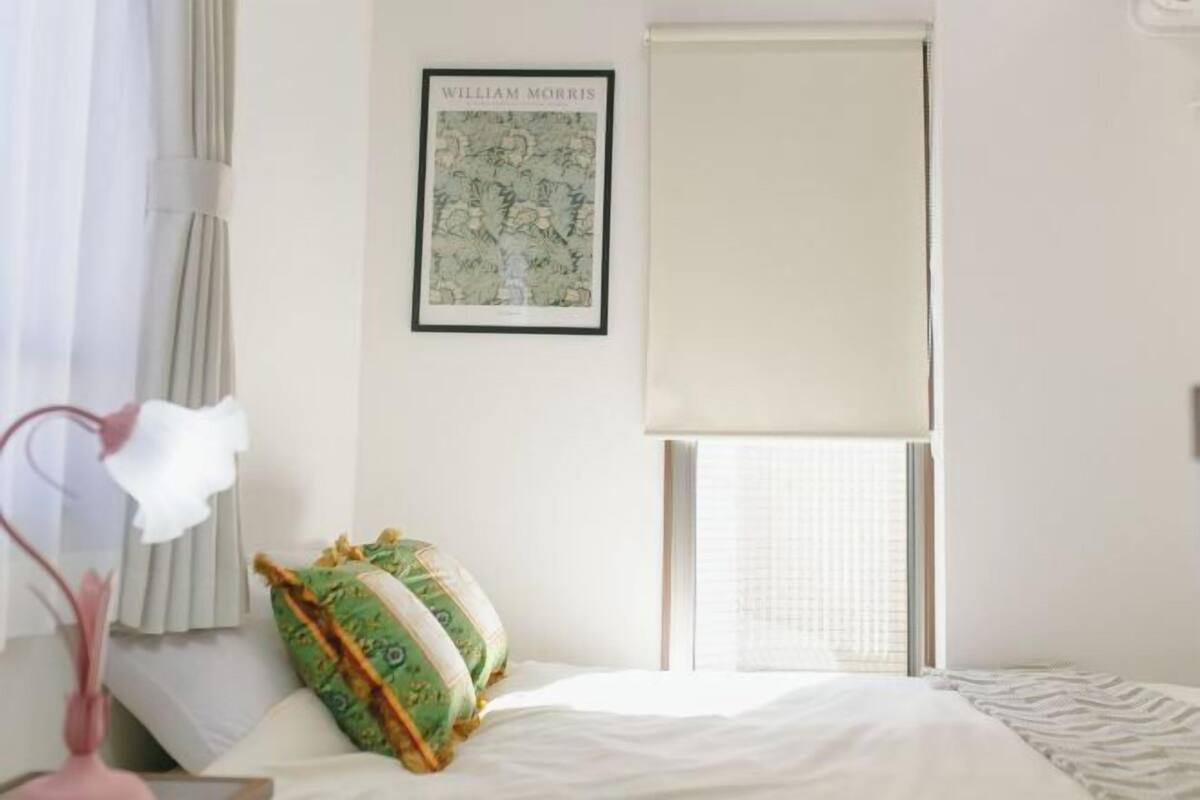 A bedroom with a bed, a framed picture on the wall, a window with blinds, and a vase with flowers.